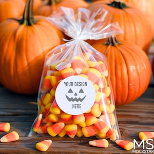 May include: A clear plastic bag filled with yellow and orange candy corn. The bag has a white circle sticker with a black jack-o'-lantern face and the text "YOUR DESIGN HERE". The bag is sitting on a wooden surface with several pumpkins in the background.