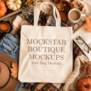 May include: A cream-colored canvas tote bag with two shoulder straps, featuring the dark brown printed text MOCKSTAR BOUTIQUE MOCKUPS Tote Bag Mockup centered on the front surface of the bag in a clear, serif font style.