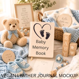 May include: A beige vegan leather baby memory book featuring a black heart and footprint graphic with the text Baby Memory Book. The book is surrounded by a tan teddy bear with a blue gingham bow, light blue crochet booties, and wooden blocks.