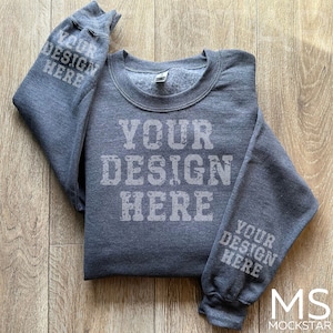 May include: A dark gray sweatshirt with the text "YOUR DESIGN HERE" printed on the front and sleeves. The sweatshirt is folded on a light brown wooden surface. The sleeves are folded over the front of the sweatshirt.