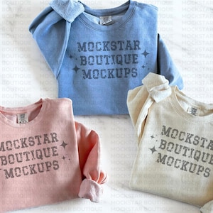 Op de afbeelding: Drie sweatshirts in verschillende kleuren: lichtblauw, roze en beige. Elk sweatshirt heeft de tekst "MOCKSTAR BOUTIQUE MOCKUPS" op de voorkant gedrukt.