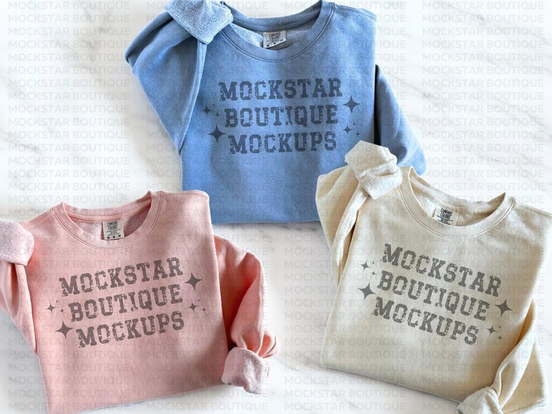 Op de afbeelding: Drie sweatshirts in verschillende kleuren: lichtblauw, roze en beige. Elk sweatshirt heeft de tekst "MOCKSTAR BOUTIQUE MOCKUPS" op de voorkant gedrukt.