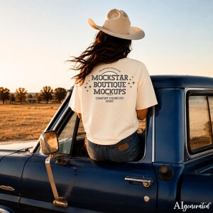 May include: A cream-colored t-shirt with the text "MOCKSTAR BOUTIQUE MOCKUPS" is worn by a person sitting on a blue vintage truck. The shirt also has the text "COMFORT COLORS 1717 IVORY". The person is wearing a beige cowboy hat.