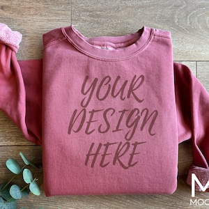 Może przedstawiać: Bordowy sweter z nadrukiem "your design here" na przodzie. Sweter leży płasko na drewnianej powierzchni.