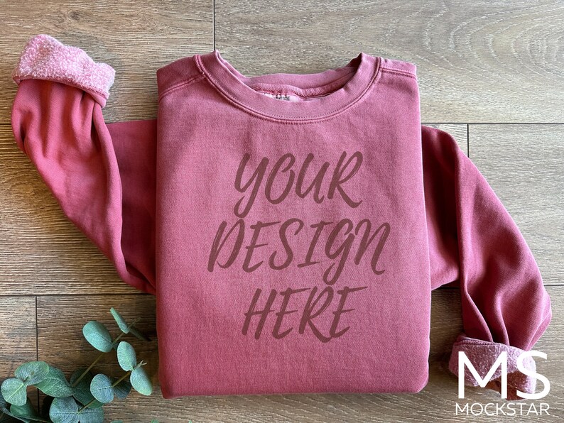 Może przedstawiać: Bordowy sweter z nadrukiem "your design here" na przodzie. Sweter leży płasko na drewnianej powierzchni.