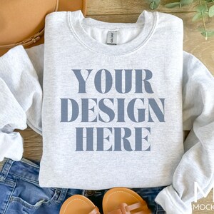 Puede incluir: Una sudadera gris con el texto "YOUR DESIGN HERE" impreso en azul en la parte delantera. La sudadera est&aacute; colocada sobre una superficie de madera.