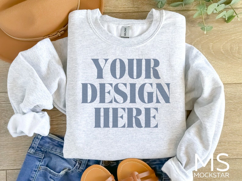 Puede incluir: Una sudadera gris con el texto "YOUR DESIGN HERE" impreso en azul en la parte delantera. La sudadera est&aacute; colocada sobre una superficie de madera.