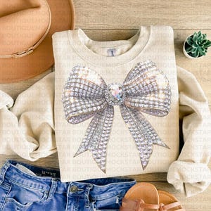 May include: A beige sweatshirt with a large, sparkly silver bow design. The bow is made of rhinestones and has a large, clear rhinestone in the center.