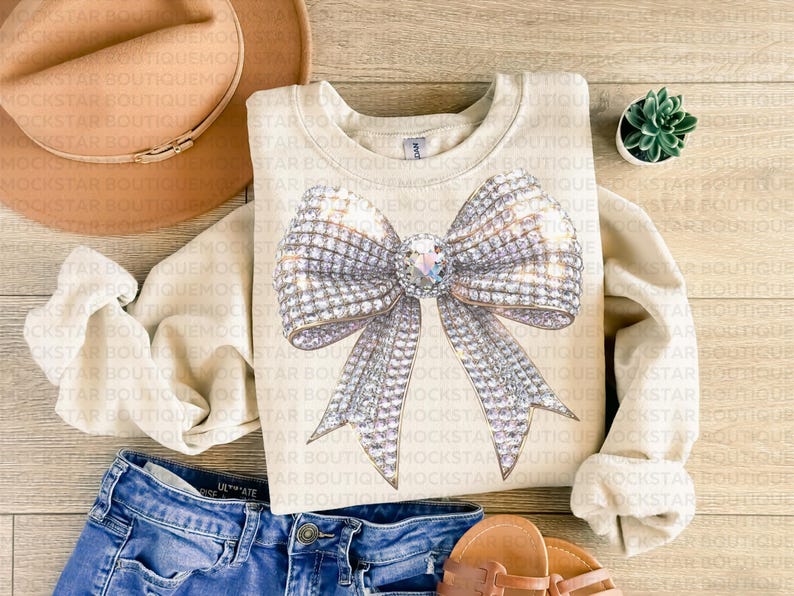May include: A beige sweatshirt with a large, sparkly silver bow design. The bow is made of rhinestones and has a large, clear rhinestone in the center.