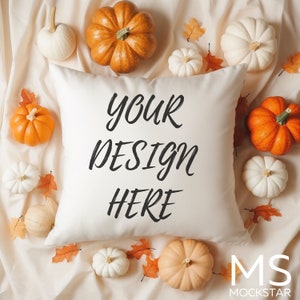 May include: A white pillow with the text "your design here" in black cursive font. The pillow is surrounded by small white and orange pumpkins and fall leaves on a cream-colored fabric background.
