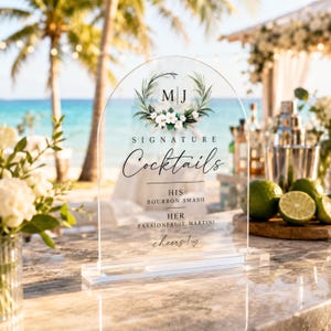 May include: An arched clear acrylic sign featuring a floral wreath graphic, the initials M J, and the text Signature Cocktails, His Bourbon Smash, Her Passionfruit Martini, and cheers! with a heart symbol, set on a rectangular base.