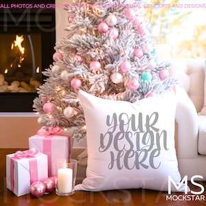 Op de afbeelding: Een wit kussen met de tekst "Your Design Here" in grijze letters. Het kussen ligt op een witte bank voor een kerstboom die versierd is met roze en witte kerstballen. Er staan ingepakte cadeaus en een kaars op de tafel voor de bank.