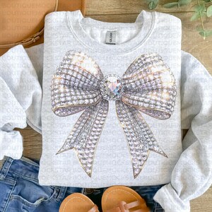 May include: A gray sweatshirt with a large, sparkly, silver bow design. The bow is made of rhinestones and has a large, clear rhinestone in the center.
