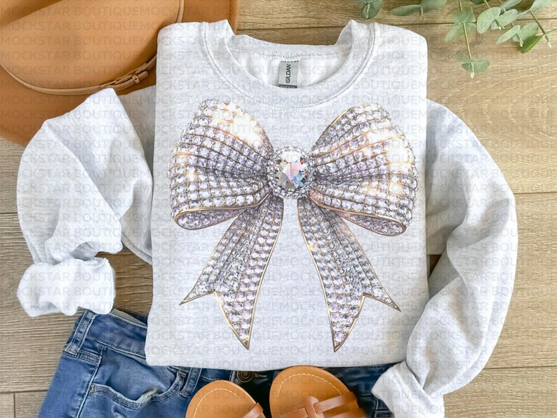May include: A gray sweatshirt with a large, sparkly, silver bow design. The bow is made of rhinestones and has a large, clear rhinestone in the center.