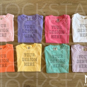 May include: Eight folded t-shirts in various colors, including pink, purple, red, white, orange, yellow, green, and lavender. Each shirt has the text "YOUR DESIGN HERE" printed on the front.
