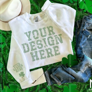 May include: White sweatshirt with the text "YOUR DESIGN HERE" in green on a green sequin background. The sweatshirt has a white hat, blue jeans, and a white clutch purse next to it.