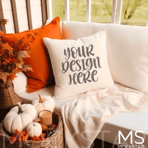 Puede incluir: Una almohada blanca con el texto "your design here" en letra cursiva gris. La almohada está en un sofá blanco con una manta blanca y almohadas naranjas. Hay decoraciones de otoño en el fondo.