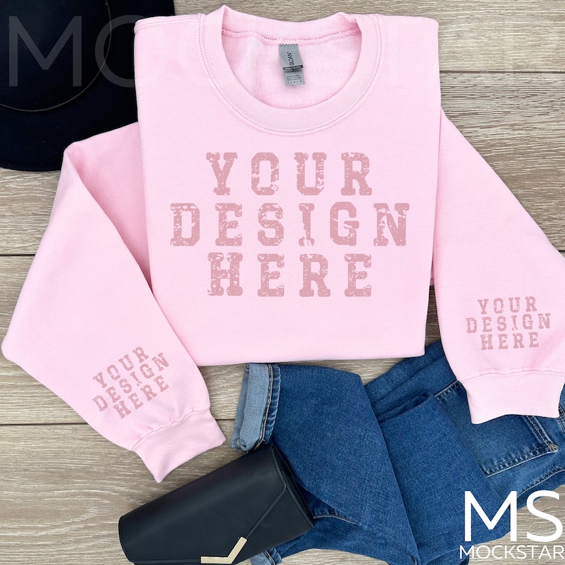 May include: Pink sweatshirt with the text "YOUR DESIGN HERE" on the front and "YOUR DESIGN HERE" on the sleeves. The sweatshirt is laid out on a wooden surface with a pair of blue jeans and a black purse.