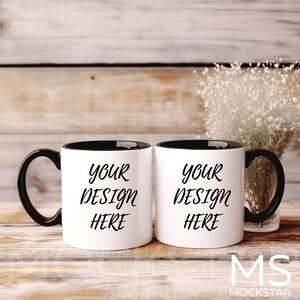 May include: Two white ceramic mugs with black handles and black rims. The mugs are on a wooden surface and both have the text "Your Design Here" printed in black.