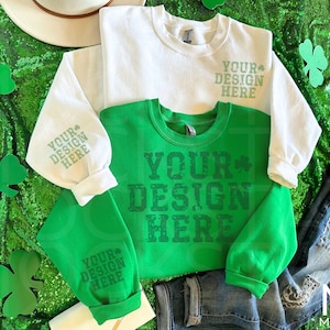 Może przedstawiać: Zielona bluza z nadrukiem "YOUR DESIGN HERE" w stylu vintage. Bluza leży na zielonym materiale z cekinami i wycięciami w kształcie koniczyny. W obrazie znajduje się również biała koszula z długim rękawem z tym samym nadrukiem.