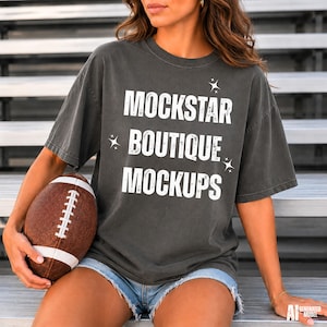 May include: A charcoal gray short-sleeve t-shirt featuring the white distressed text MOCKSTAR BOUTIQUE MOCKUPS with small star icons. The shirt is paired with blue denim shorts and a brown leather football with white laces held in one hand.