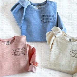 Op de afbeelding: Drie ronde hals sweaters in verschillende kleuren: lichtblauw, roze en beige. Elke sweater heeft de tekst "MOCKSTAR BOUTIQUE MOCKUPS" op de voorkant gedrukt.