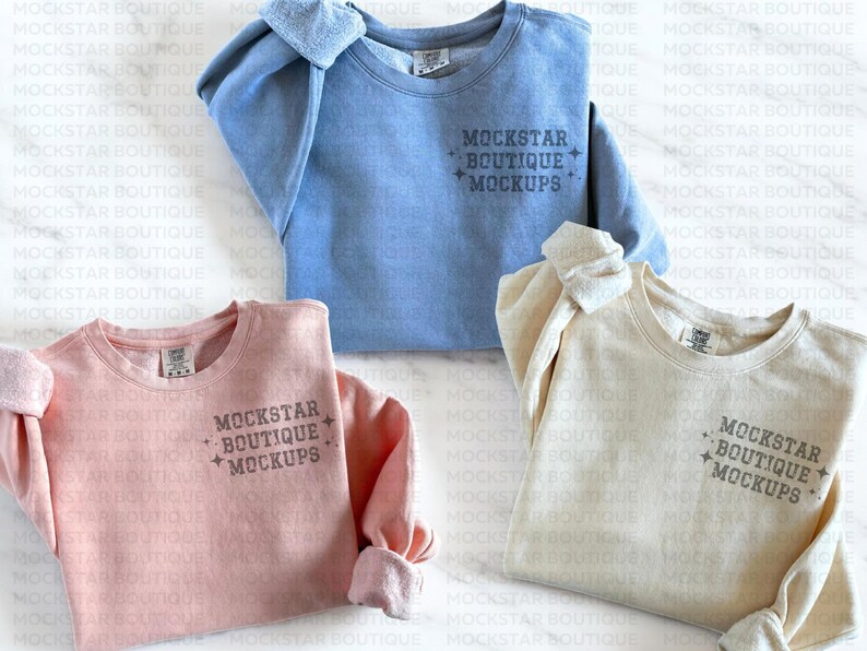 Op de afbeelding: Drie ronde hals sweaters in verschillende kleuren: lichtblauw, roze en beige. Elke sweater heeft de tekst "MOCKSTAR BOUTIQUE MOCKUPS" op de voorkant gedrukt.