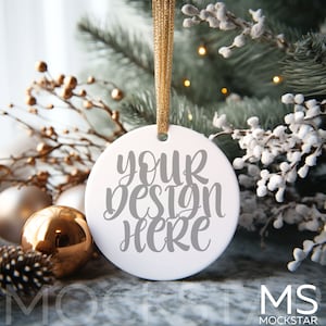 May include: White ceramic ornament with a gold string hanging from a snowy pine branch. The ornament has the text "your design here" written in gray.
