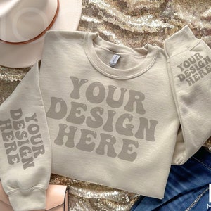 Könnte beinhalten: Ein beiger Pullover mit dem Text "YOUR DESIGN HERE" in einer Retro-Schriftart auf der Vorderseite und den Ärmeln. Der Pullover liegt auf einem goldenen Paillettenstoff.