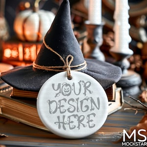 May include: A black witch hat with a decorative twine tie rests on a stack of old books. A white, round ornament with the text "YOUR DESIGN HERE" hangs from the hat, perfect for Halloween decor.