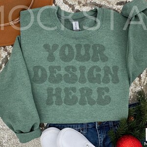 May include: A green sweatshirt with the text "YOUR DESIGN HERE" in a retro font. The sweatshirt is laying on a sparkly surface with a brown hat and a sprig of greenery.