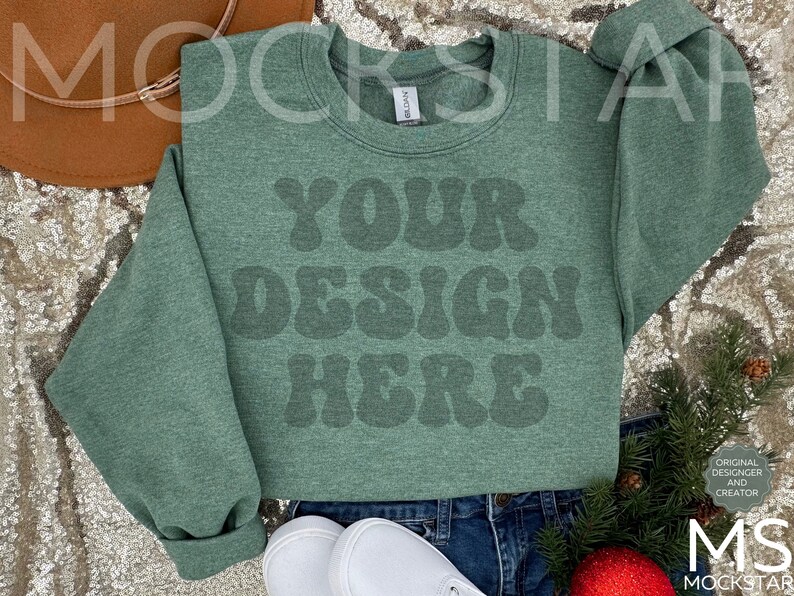 May include: A green sweatshirt with the text "YOUR DESIGN HERE" in a retro font. The sweatshirt is laying on a sparkly surface with a brown hat and a sprig of greenery.