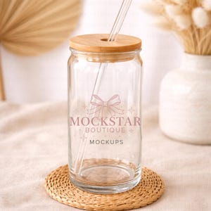 May include: A clear glass tumbler with a wooden lid and a glass straw. The tumbler has a pink bow and the text "MOCKSTAR BOUTIQUE MOCKUPS" printed on the front. It sits on a round, woven coaster.