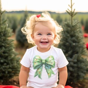 May include: A young child wearing a white onesie with a green Christmas tree bow with lights on it. The child is smiling and looking up. The child is in a field of Christmas trees.