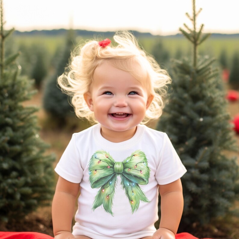 May include: A young child wearing a white onesie with a green Christmas tree bow with lights on it. The child is smiling and looking up. The child is in a field of Christmas trees.