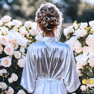 May include: A white satin robe with the text "your design here" printed on the back. The robe is worn by a person with their back to the camera, standing in front of a wall of white roses.