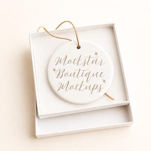 May include: A round white ceramic ornament featuring the brown text Mockstar Boutique Mockups with small star accents. The ornament includes a gold hanging string and is presented inside an open white cardboard gift box with a soft lining.