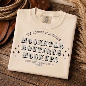May include: Ivory-colored t-shirt with the text "THE MOCKUP COLLECTION MOCKSTAR BOUTIQUE MOCKUPS" in black. The shirt is folded and placed on a wooden surface. The tag reads "COMFORT COLORS XL-TG-EG".
