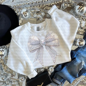 May include: A white sweatshirt with a large sparkly silver bow design. The sweatshirt is laying on a silver sequin fabric with a black hat, blue jeans, and a black purse.