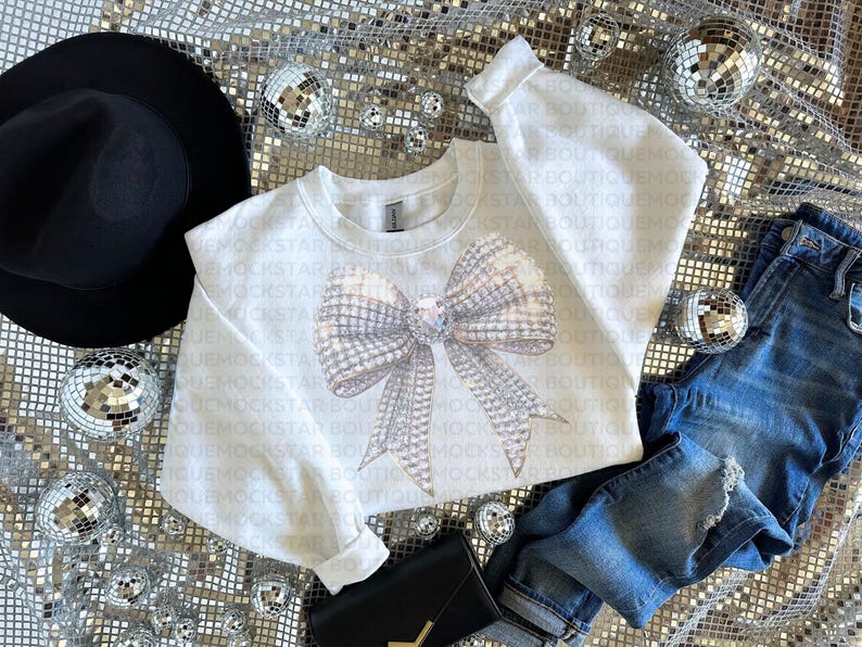 May include: A white sweatshirt with a large sparkly silver bow design. The sweatshirt is laying on a silver sequin fabric with a black hat, blue jeans, and a black purse.
