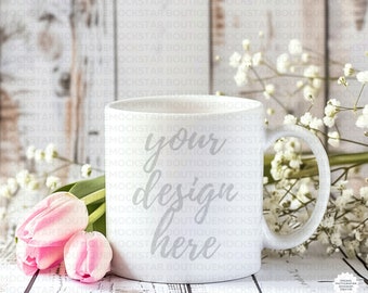 Könnte beinhalten: Weiße Keramik-Tasse mit dem Text "your design here" in Schreibschrift. Neben der Tasse liegen rosa Tulpen und weiße Blüten auf einem rustikalen Holzgrund.
