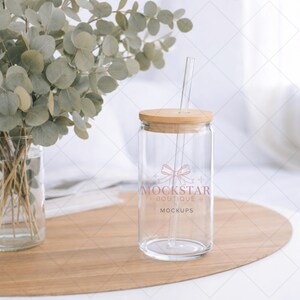 May include: A clear glass tumbler with a wooden lid and a clear straw. The tumbler has the text "MOCKSTAR BOUTIQUE MOCKUPS" printed on it. The tumbler is on a wooden tray with a vase of green leaves in the background.