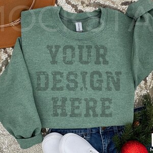May include: A green sweatshirt with the text "YOUR DESIGN HERE" printed on the front. The sweatshirt is laying on a sparkly surface with a brown hat and a sprig of greenery.
