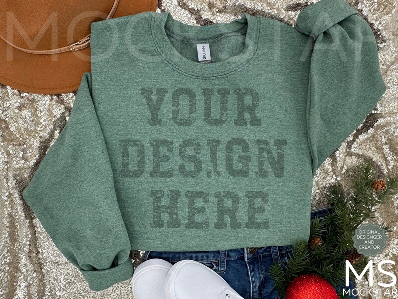 May include: A green sweatshirt with the text "YOUR DESIGN HERE" printed on the front. The sweatshirt is laying on a sparkly surface with a brown hat and a sprig of greenery.