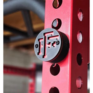 May include: A black and red weightlifting rack accessory. The round black piece has a textured edge and a cut-out design with the letters "FF". The rack is red with several round holes.