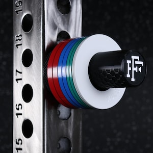 May include: A black, white, green, blue, and red weight plate storage pin attached to a vertical steel rack with numbered holes. The pin features a black end cap with a white logo displaying the letters FF in a stylized, interlocking font.