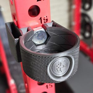 May include: Black metal cup holder with a textured surface and a logo. The cup holder is attached to a red metal pole.