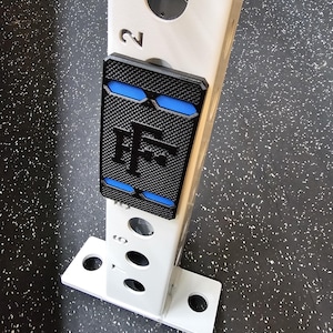May include: A black and blue textured plate with the letters "TF" is attached to a white metal support structure. The support structure has a series of holes and the number "2" is visible. The background is a speckled black and gray surface.