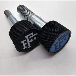 May include: Two metal bolts with textured black knobs. Each knob has an oval-shaped insert with the white letters "FF" on a black or blue background. The bolts are on a white surface.