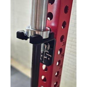 Può includere: Un porta bilanciere in metallo nero fissato a un rack di pesi in metallo rosso. Il porta bilanciere ha un clip in plastica nera e una vite in metallo. Il rack ha una scala di numeri bianca sul lato.